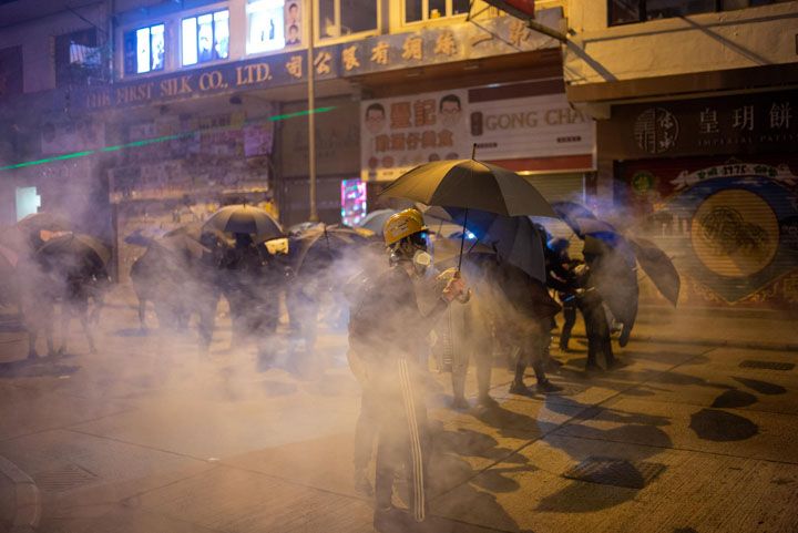 people in helmets carrying umbrellas in a cloud of tear gas